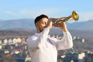 Trubač, Aleksandar Stojanović, u beloj tradicionalnoj košulji sa vezom svira trubu sa zatvorenim očima, dok se u pozadini pruža pogled na Vladičin Han i planine.