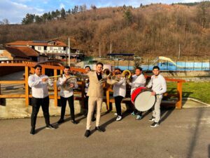 Muzičari iz Orkestra AS stoje na mostiću na otvorenom, obučeni u bela i bež odela, držeći trube, doboš i ostale instrumente, spremni za svirku.